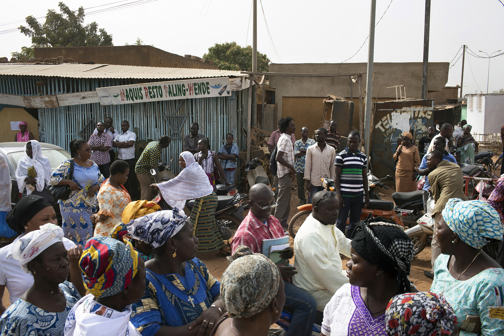 Scène de rue : des piétons crosient des personnes en scooter sur une piste qui passe au ras d'échoppes, Ouagadougou, Burkina Faso, 2016.