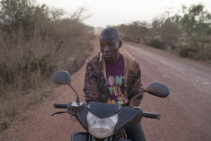 Portrait d'un jeune homme assis sur son scooter, couvert de latérite après un voyage au village de Manevire, à 30 km de Ouagadougou, Burkina-Faso, 2016.