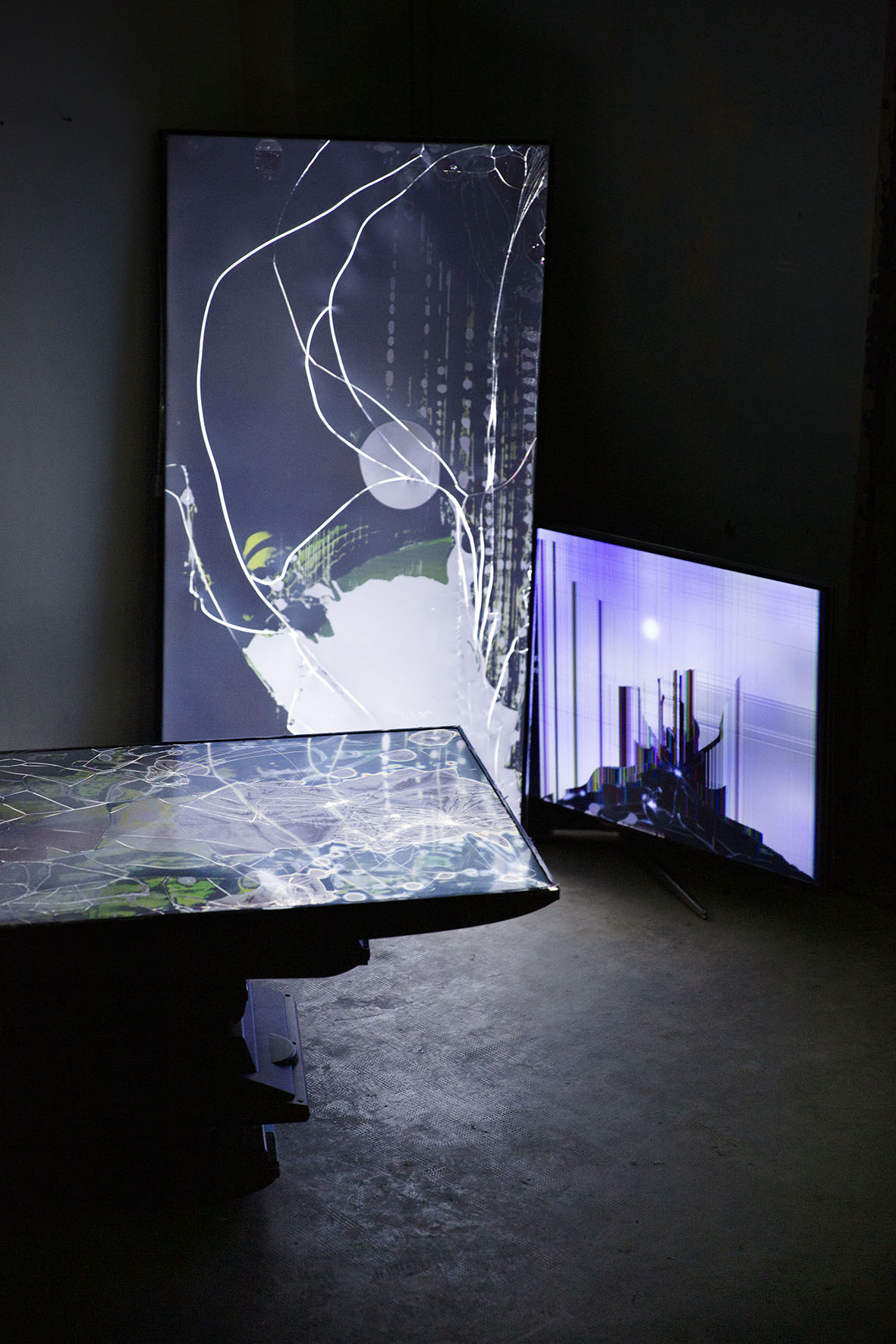 Plan rapproché d'une installation en volume : dans une pièce sombre, des écrans défectueux mis sous tension donnent à voir des traces d'impact et des effets picturaux.